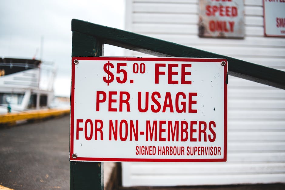 A white rectangular sign with red text displaying a fee of £5.00 per usage for non-members, attached with four screws to a dark green metal frame. The sign is positioned outdoors, near a dock or quay