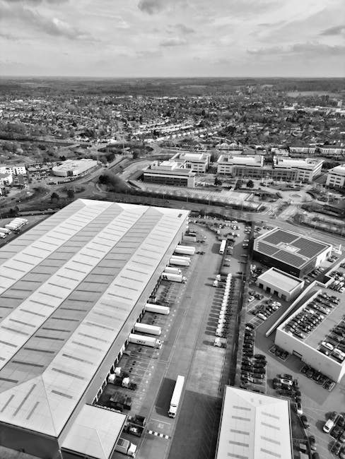 An aerial view in black and white of a large commercial warehouse complex and surrounding parking lot, featuring multiple loading bays with several trucks and delivery vans parked adjacent to the building. In the foreground, a long, rectangular warehouse with a corrugated metal roof has multiple dock doors aligned along its side, with some trucks actively positioned for loading or unloading. The parking area is partially filled with cars and additional service vehicles, with clear designated lanes and pedestrian pathways. Beyond the warehouse and parking lot, the image shows an expansive urban landscape with office buildings, roads, and residential areas extending into the distance under a partly cloudy sky. This scene highlights the logistics involved in home relocation and furniture transport, illustrating the loading process and the infrastructure supporting moving services, as managed by Man with Van Barnsbury.