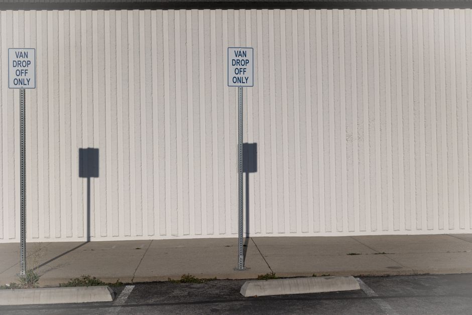 Photograph of two blue and white van drop-off only parking signs mounted on metal posts along a paved pavement in front of a white corrugated metal wall. The signs are positioned next to each other, with the left sign slightly to the left and the right sign centered in the image. Shadows cast by the signs fall onto the wall, and the pavement below shows minor vegetation growth at the edge. The scene captures a designated unloading zone typically used for household furniture transport and home relocation services, with no vehicles present. The setting suggests an area reserved for moving company vehicles, such as Man with Van Barnsbury, to park temporarily for loading or unloading household goods, boxes, furniture, or packaging materials during a house move or relocation process.