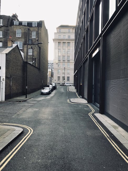 A narrow urban street scene showing a residential and commercial area with parked cars along the curb, including a small white van positioned near a modern black building with large window panels and a metal roll-up door. The street has double yellow lines indicating parking restrictions and slight road markings at the edge. In the background, taller older brick buildings with multiple windows are visible, and the sky appears overcast with diffuse lighting. This setting may be part of a typical home relocation or furniture transport process, where vehicles are parked close to the property for loading or unloading boxes, furniture, and packing materials as part of an effective removals service, such as those provided by Man with Van Barnsbury, working within the constraints of narrow streets and parking regulations.