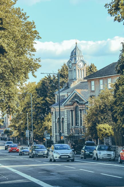A row of parked cars along a street on a bright day, with a historic clock tower building featuring ornate architecture, arched windows, and a spire visible behind leafy trees. The street is lined with residential and commercial buildings, with some facing the pavement and others set back behind greenery. The scene includes a clear sky with a few clouds, and street lighting poles along the pavement. In the context of house removals and transport, Man with Van Barnsbury's services assist with home relocation logistics, including moving furniture and boxes through urban environments like this one, where narrow streets and parking considerations are important for an efficient loading process.