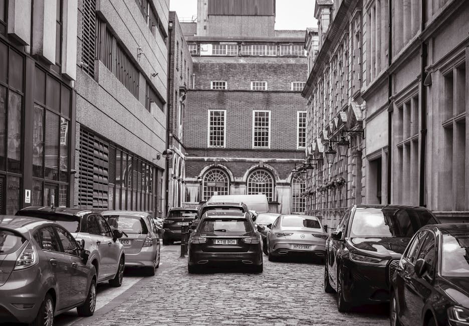 A narrow urban street scene showing a residential and commercial area with parked cars along the curb, including a small white van positioned near a modern black building with large window panels and a metal roll-up door. The street has double yellow lines indicating parking restrictions and slight road markings at the edge. In the background, taller older brick buildings with multiple windows are visible, and the sky appears overcast with diffuse lighting. This setting may be part of a typical home relocation or furniture transport process, where vehicles are parked close to the property for loading or unloading boxes, furniture, and packing materials as part of an effective removals service, such as those provided by Man with Van Barnsbury, working within the constraints of narrow streets and parking regulations.