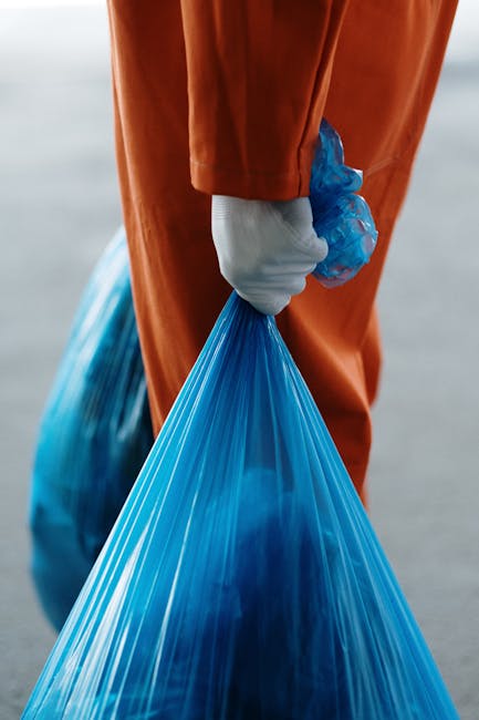 A close-up view of a person wearing an orange safety suit and white gloves, holding a large blue plastic rubbish bag filled with waste. The individual is standing outdoors on a paved surface, with part of their leg and hand visible, and the background appears blurred, indicating an active waste collection or clearance process. This scene relates to waste removal, which can be a part of house and furniture clearance services offered by Man with Van Barnsbury as part of their remit in removals and home relocation logistics. The focus on protective clothing and waste containment highlights the disposal aspect of moving or clearance projects, supporting efficient packing and loading procedures during furniture transport or house packing tasks.