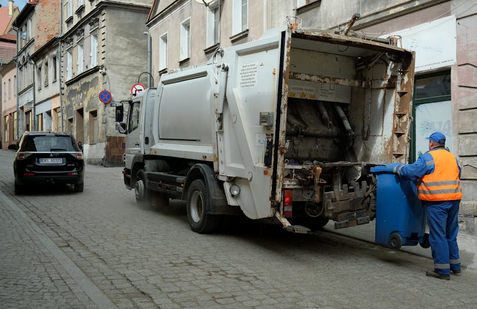 A close-up view of a person wearing an orange safety suit and white gloves, holding a large blue plastic rubbish bag filled with waste. The individual is standing outdoors on a paved surface, with part of their leg and hand visible, and the background appears blurred, indicating an active waste collection or clearance process. This scene relates to waste removal, which can be a part of house and furniture clearance services offered by Man with Van Barnsbury as part of their remit in removals and home relocation logistics. The focus on protective clothing and waste containment highlights the disposal aspect of moving or clearance projects, supporting efficient packing and loading procedures during furniture transport or house packing tasks.