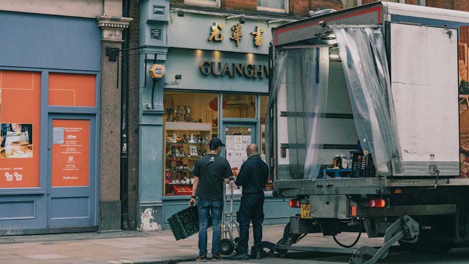 Two movers from Man with Van Barnsbury are preparing a house relocation outside a shop named Guangh, which features a grey facade with gold Chinese characters and the word 'GUANGH' in large metal lett