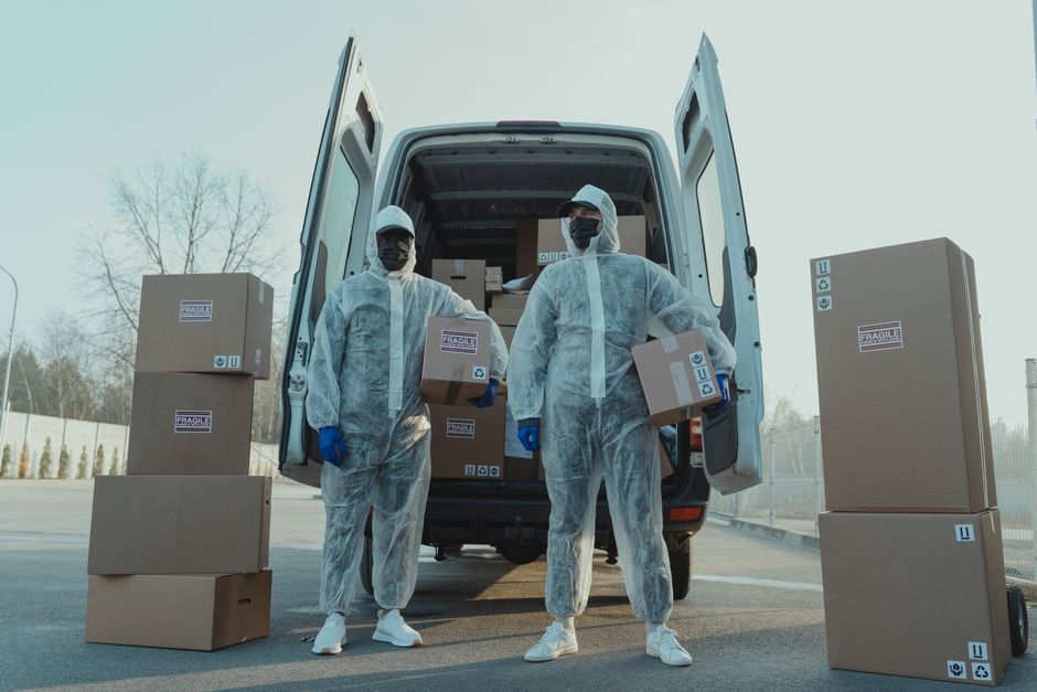 Two movers dressed in full protective coveralls, face masks, and gloves are standing outdoors in front of an open van, which is parked on a paved area. The van's interior is visible, showing packed ca
