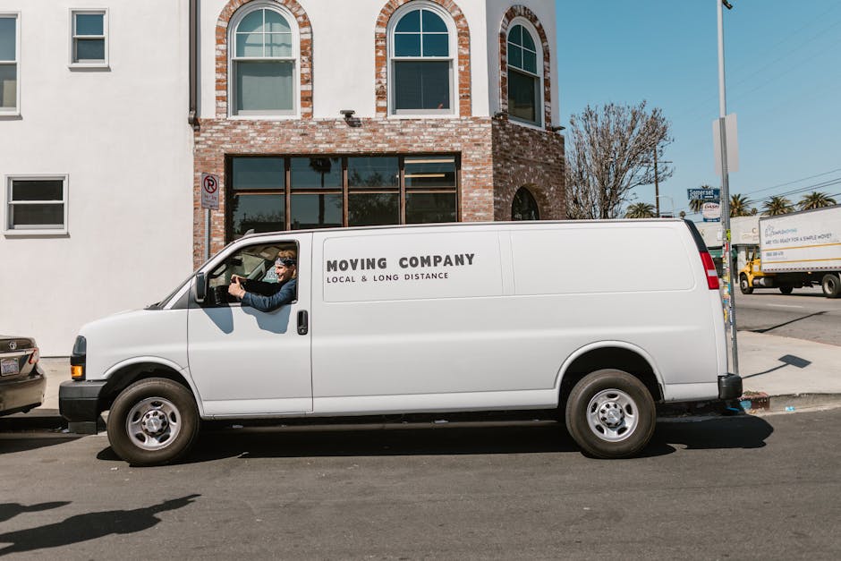 Two movers dressed in full protective coveralls, face masks, and gloves are standing outdoors in front of an open van, which is parked on a paved area. The van's interior is visible, showing packed ca