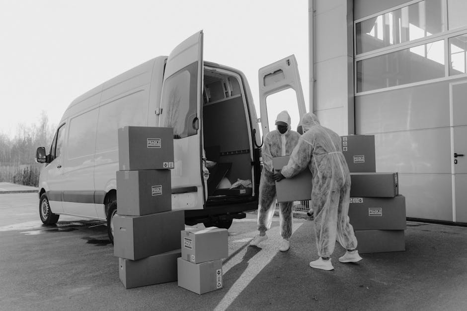 Two movers dressed in protective overalls, face masks, and hair coverings are loading cardboard boxes onto a white panel van arranged outside a modern building with large glass windows. The boxes vary in size and are stacked on the ground and partially on the van’s open rear doors, which reveal an empty cargo space. The scene is set on a paved area with a clear sky and some trees visible in the background. The movers are carefully handling the boxes, with some already inside or about to be placed into the van as part of a home relocation process. This image illustrates furniture transport and packing activities typical of a professional removals service, supported by the presence of a moving van and packaging materials, aligning with the services offered by Man with Van Barnsbury for house moves and relocation logistics.