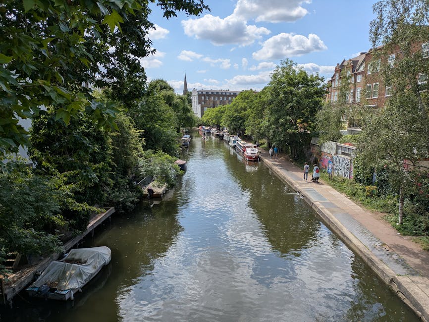 A wide view of a narrow, calm canal lined with lush green trees on both sides, with several boats docked along the water's edge. On the right side, a paved footpath runs parallel to the canal, with a few pedestrians walking and some residential buildings with brick facades and bay windows visible behind the trees. Parked cars are also seen along the street adjacent to the path. The skyline features a partly cloudy sky with scattered white clouds and a tall church spire in the distance. This setting depicts an urban residential area suitable for house removals and home relocation projects, with a focus on the outdoor environment and street-side activities often associated with moving logistics, as managed by Man with Van Barnsbury, specialists in removals. The scene emphasizes the peaceful streetscape and the connecting waterways characteristic of Barnsbury and Islington, relevant for transporting furniture and belongings during a move.