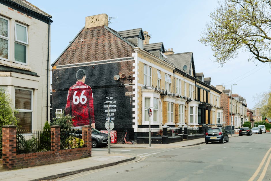 A mural painted on the side of a brick building on Liverpool Road depicts a man in a red sports jersey with the number 66 and the name Alexander-Arnold on the back, facing away from the viewer. The mural includes white lettering underneath the figure, with some text partially obscured. The building is part of a row of Victorian terraced homes with bay windows, decorative iron balconies, and varied brickwork, situated on a tree-lined street with parked cars along the pavement. A small front garden with a brick wall and metal fencing surrounds one property, adjacent to the mural. The street has a traffic sign, a scooter parked near the curb, and a street lamp. The scene takes place during daylight with overcast sky, capturing the environment where a professional moving service like Man with Van Barnsbury could be involved in home relocation or furniture transport, evidenced by the presence of parked vehicles and urban setting.