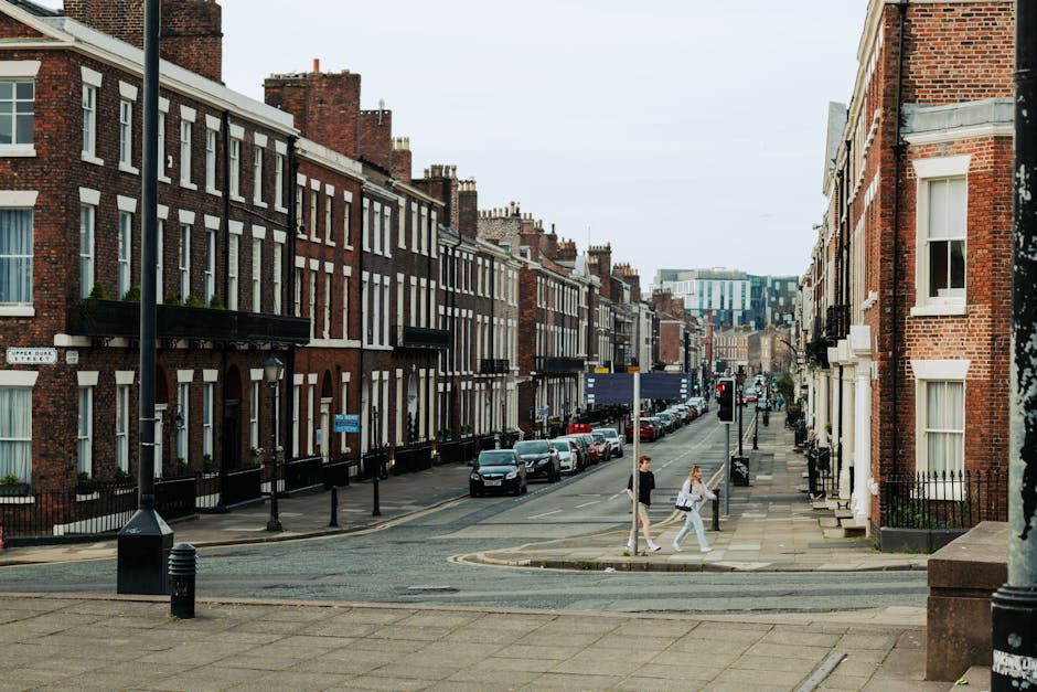 The image shows a typical residential street scene in a Victorian terraced neighbourhood with rows of red-brick houses featuring white-framed windows and small front gardens with black wrought-iron fences. Several parked cars line both sides of the narrow street, which is paved with asphalt. Two pedestrians, a man and a woman, are crossing the sidewalk near the curb, each carrying bags, indicating a typical day in the area. The street is lit by tall lampposts, and additional black bollards separate the pavement from the road. The environment appears well-maintained with traditional architecture, and the lighting suggests daytime. This scene accurately reflects a typical UK city street suitable for house removals or home relocation logistics, with potential for furniture transport and moving activities as handled by companies like Man with Van Barnsbury.