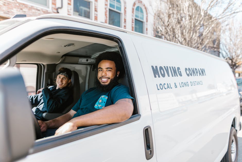 Inside a white moving van operated by Man with Van Barnsbury, a professional removals company, two movers are visible preparing for home relocation near Union Chapel. The driver, a man with a beard and dark curly hair, is smiling at the camera, wearing a navy blue uniform with a blue logo, sitting behind the steering wheel. In the passenger seat, a woman with dark hair, dressed in a black jacket and a headband, is also seated. The van's interior shows various packing materials including cardboard boxes, plastic-wrapped furniture, and blankets for protection during furniture transport. The van is parked on the street with the side door open, revealing the loading process for furniture and boxes. The background includes a residential building, trees without leaves, and a clear sky, indicating a daytime setting. This scene demonstrates the logistics involved in packing, loading, and organizing belongings during a professional move, as part of the services offered by Man with Van Barnsbury in the context of house removals near Union Chapel.
