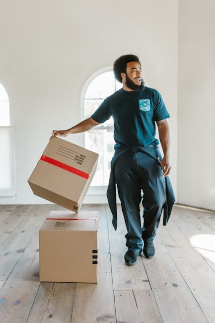 A man with a dark beard and curly hair, wearing a blue T-shirt with a logo on the chest and dark trousers, is seen indoors during a home relocation. He is lifting one cardboard box with red tape around it, placing it on top of another box on a light wooden floor. The room has white walls and features a large arched window allowing natural daylight to illuminate the space. In the background, there are no additional furniture or appliances visible, emphasizing the process of packing and loading during furniture transport or a house move. The scene captures the careful handling of boxes as part of a professional removals service, aligned with the moving near Union Chapel theme, and reflects the importance of organized packing and safe loading in house removals, with [COMPANY_NAME] providing expert moving support. The overall environment appears clean and minimal, suitable for professional packing and furniture transport operations as part of a detailed house relocation process.