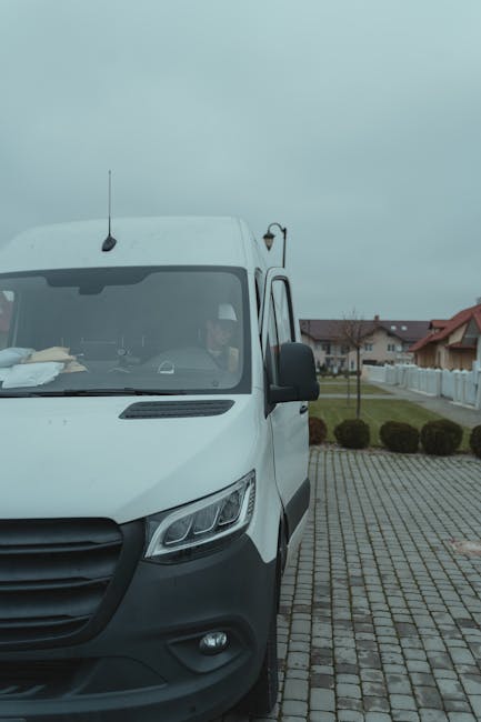 A young man with light brown hair, dressed in a blue hoodie, navy trousers, and white sneakers, is sitting on the edge of an open moving van with an interior composed of metal panels and insulation, s