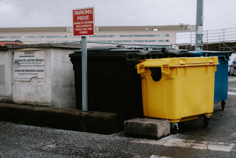 A row of three large wheelie bins positioned on a city street pavement adjacent to a concrete wall with signage. The first bin is black, the second bright yellow, and the third blue, all with closed lids and small caster wheels at the base. The yellow bin has a black latch on the front, and the bins are located next to a concrete curb with a small concrete block placed in front of the yellow bin to stabilize it. Behind the bins, there is a red and white parking sign attached to a metal pole, indicating a designated parking area. The background shows an urban environment with modern, multi-story buildings and a clear sky, suggesting a typical residential or commercial setting. This scene illustrates typical waste management or clearance setup, relevant to home relocation or packing and moving tasks performed by companies like Man with Van Barnsbury, involved in removals and furniture transport services.