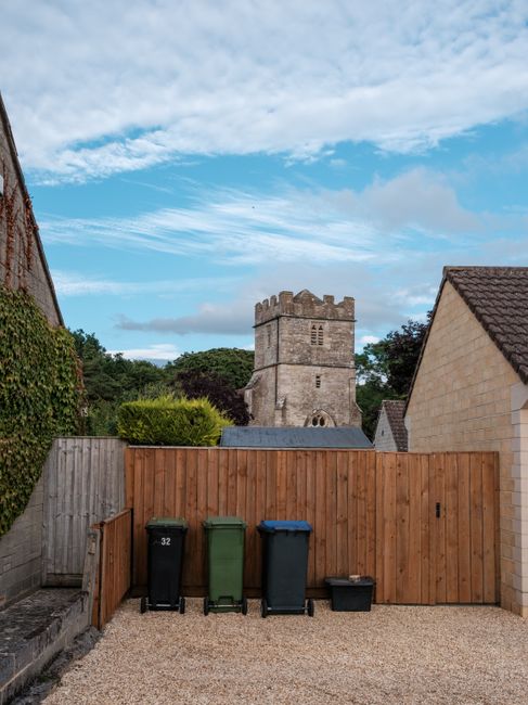 A residential backyard area showing a wooden fence with four wheeled waste bins in front — two black, one green, and one blue — positioned on a gravel surface. To the left, part of a brick house wall covered with climbing plants is visible. In the background, a historic stone tower with battlements and small windows rises above trees and greenery, indicating a rural setting. The sky above is partly cloudy with patches of blue showing through. This outdoor scene captures a typical home exterior, relevant to the context of removals and moving logistics as part of home relocation or clearing activities, supported by the presence of waste disposal containers and the open space for loading or unloading activities, demonstrating the environment where furniture transport and packing processes may occur. Man with Van Barnsbury occasionally offers such moving services, including loading and transport within residential areas.