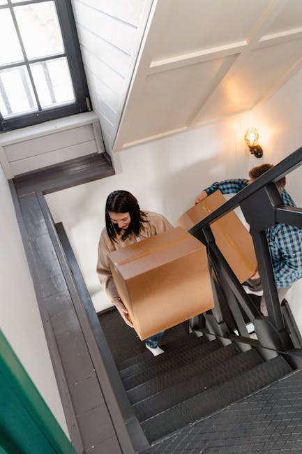 A woman with dark hair, dressed in a beige sweater, is carrying a large cardboard box down a staircase inside a residential building, assisted by a man in a blue checkered shirt. The staircase has dark carpeting with a black handrail, and the walls are painted white with a decorative light fixture on the wall. A window at the top of the stairs allows natural light into the area. The scene depicts a home relocation or furniture transport process, with the two individuals carefully navigating the staircase as part of a moving or packing and moving service provided by Man with Van Barnsbury. The staircase space is narrow, indicating challenges typical of flats with limited access, and the individuals are using proper lifting techniques and equipment, such as blankets and straps, to ensure safe furniture moving.