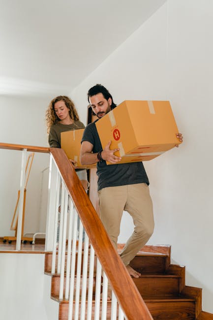 A man and a woman are carrying cardboard boxes during a home relocation on a staircase inside a residential property. The man, positioned at the front, holds a large cardboard box wrapped with packing tape, with a red label or symbol visible on the side. Behind him, the woman is also holding a similar box, both standing on wooden stairs with a wooden handrail. The staircase is inside a well-lit interior space with white walls, and the scene captures the process of furniture transport and packing during a move. The image illustrates the logistics involved in house removals, with the movers carefully navigating a narrow stairway, typical of relocation services offered by companies like Man with Van Barnsbury. Equipment such as blankets or straps is not visible, but the focus is on the safe handling of boxes during an internal moving process, consistent with professional furniture transport in urban flats like those in Barnsbury.