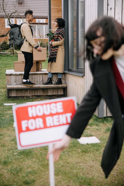 A young man wearing a beige jacket and black pants stands on a small wooden porch holding a cardboard box, engaged in conversation with a woman dressed in a striped coat and blue jeans, who is also holding a box. They are positioned outside a modern house with a glass door and window. Several cardboard boxes are stacked on the porch, indicating an active home relocation process. In the foreground, a blurred figure, likely a woman, is partially visible, holding a red and white 'House for Rent' sign, which is slightly out of focus. The scene is set on a grassy lawn with a neighboring house visible in the background. The environment appears bright and clear, capturing the logistics of packing and moving, with the team from Man with Van Barnsbury managing the transportation of furniture and belongings during an indoor-to-outdoor loading process as part of a house removal or relocation service.