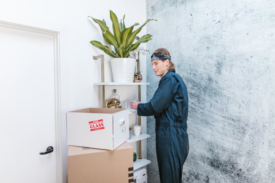 A young man wearing a beige jacket and black pants stands on a small wooden porch holding a cardboard box, engaged in conversation with a woman dressed in a striped coat and blue jeans, who is also holding a box. They are positioned outside a modern house with a glass door and window. Several cardboard boxes are stacked on the porch, indicating an active home relocation process. In the foreground, a blurred figure, likely a woman, is partially visible, holding a red and white 'House for Rent' sign, which is slightly out of focus. The scene is set on a grassy lawn with a neighboring house visible in the background. The environment appears bright and clear, capturing the logistics of packing and moving, with the team from Man with Van Barnsbury managing the transportation of furniture and belongings during an indoor-to-outdoor loading process as part of a house removal or relocation service.